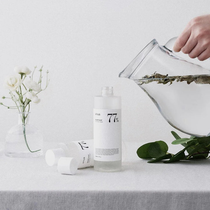 Anua - 
Heartleaf 77% Soothing Toner står på et bord med det hvide lå pillet af.. der ligger en flaske mere på bordet .. der er en blomst og er person er ved at hælde klar væske med nogle blade i en kande nede i toner beholderen