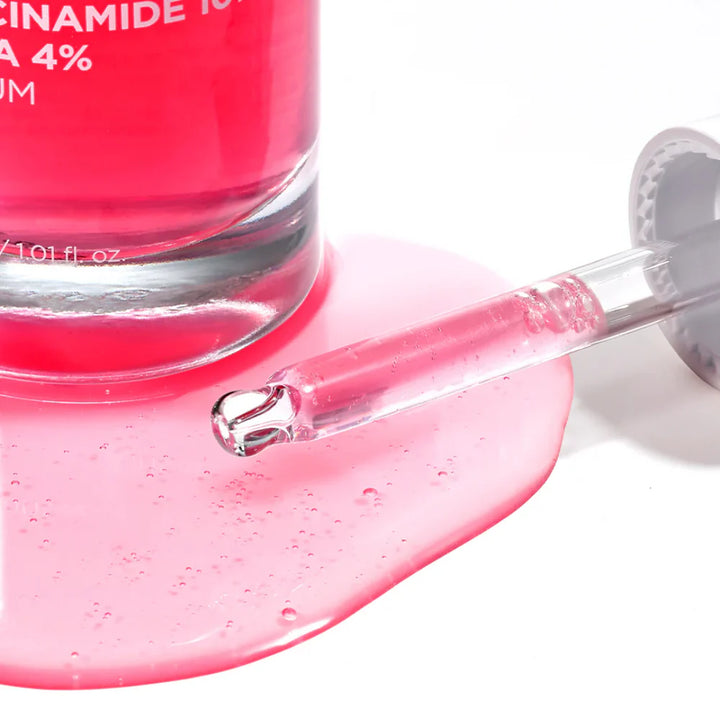 En pipette fyldt med rød væske fra Anua Niacinamide 10% + Tranexamsyre 4% Serum ligger ved siden af en delvist synlig flaske. Der er masser af rød serum synlig i både pipetten og flasken, og en dråbe rød væske er ved at falde fra pipettens spids. Baggrunden er lys.