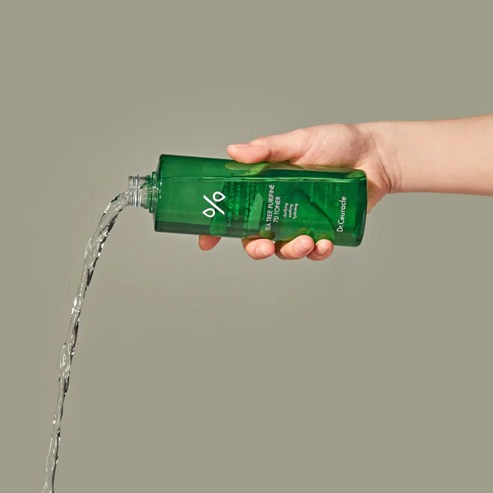 En kvinde med ren, lys hud og mørkt hår hælder forsigtigt Dr. Ceuracle Tea Tree Purfine 70 Toner fra en grøn flaske ud i sin håndflade. Hun smiler let og står i et lyst, minimalistisk miljø, hvilket understreger produktets rene og naturlige profil.