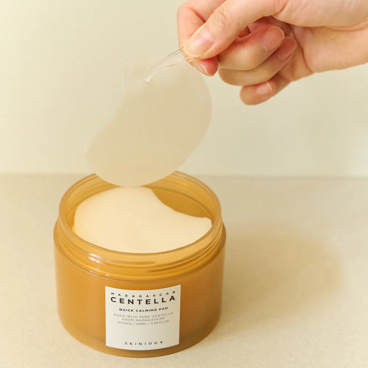 En enkelt rund, hvid pad fra SKIN1004 Madagascar Centella Quick Calming Pad e holdes op over den åbne beholder, så både pad og beholder er synlige