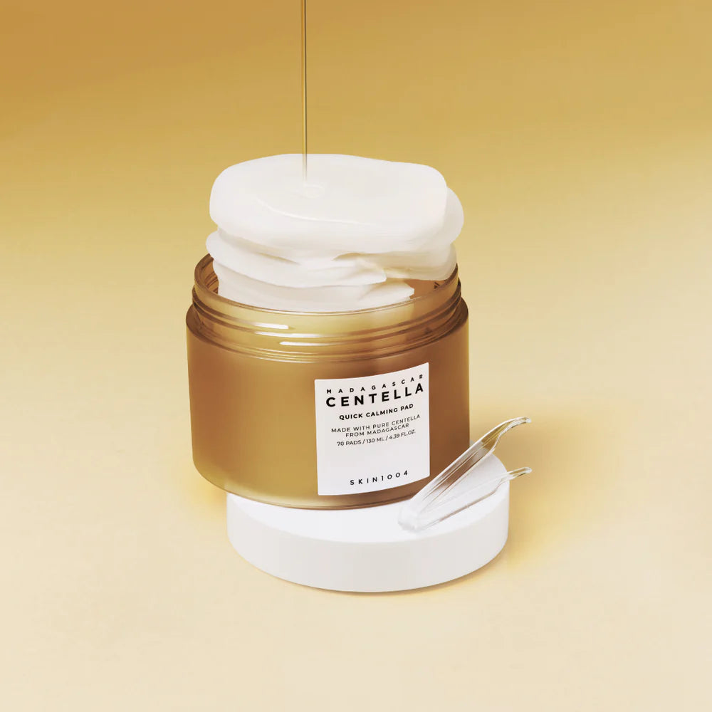 SKIN1004 Madagascar Centella Quick Calming Pad – rund beholder med hvidt låg, fotograferet fra en vinkel, så både låg og etiket er synlige. Produktet står på en gullig baggrund. man kan se noget essens blive hældt ud over nogle pads