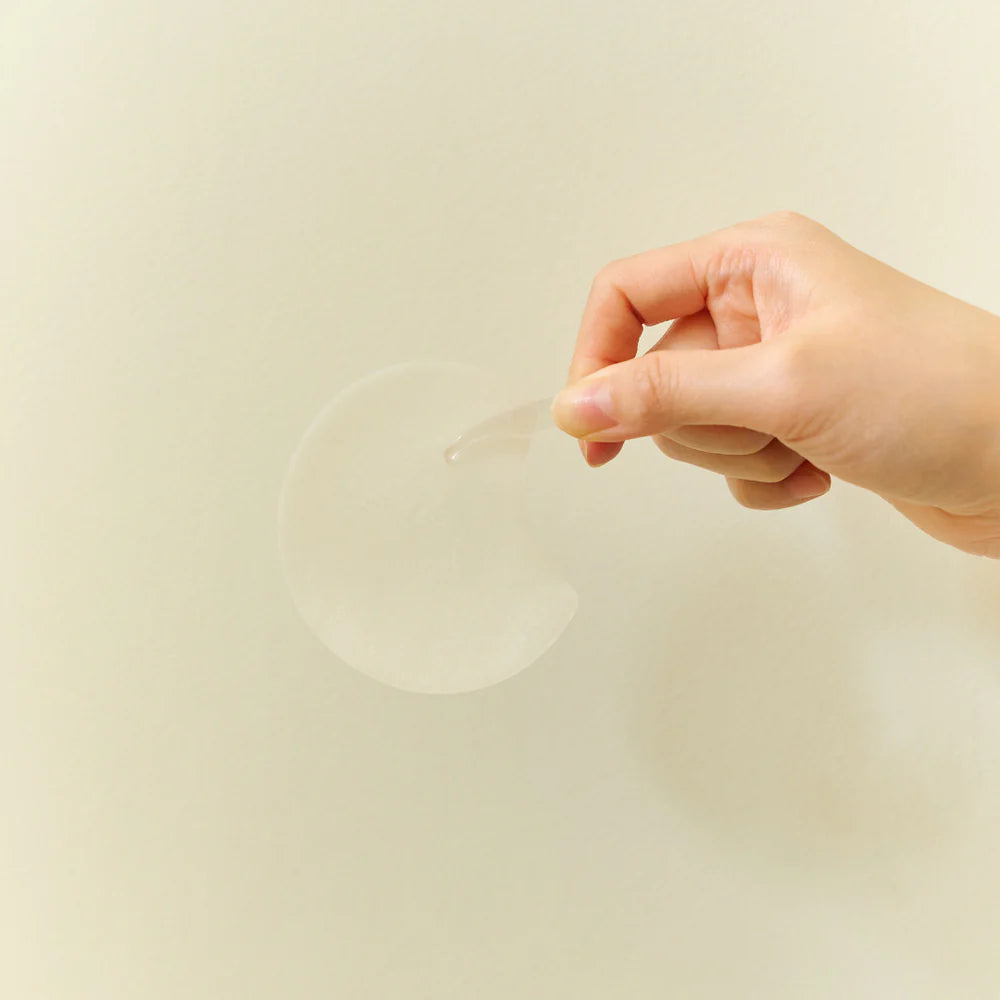 En hånd holder en enkelt rund, hvid pad fra SKIN1004 Madagascar Centella Quick Calming Pad op mod en lys baggrund. Ingen beholder er synlig på billedet.